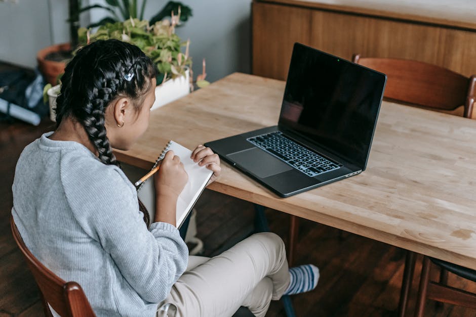 Back view of little girl taking notes in notepad while sitting at desk and watching laptop